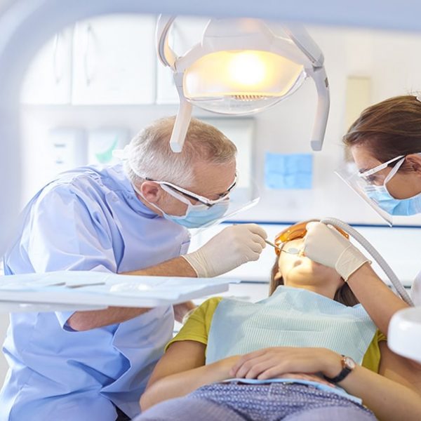 A senior dentist scrapes away some debris from between a patient tooth while the dental nurse applies suction in the mouth to take away any foreign objects. The brightly lit dentist surgery is clean and modern.